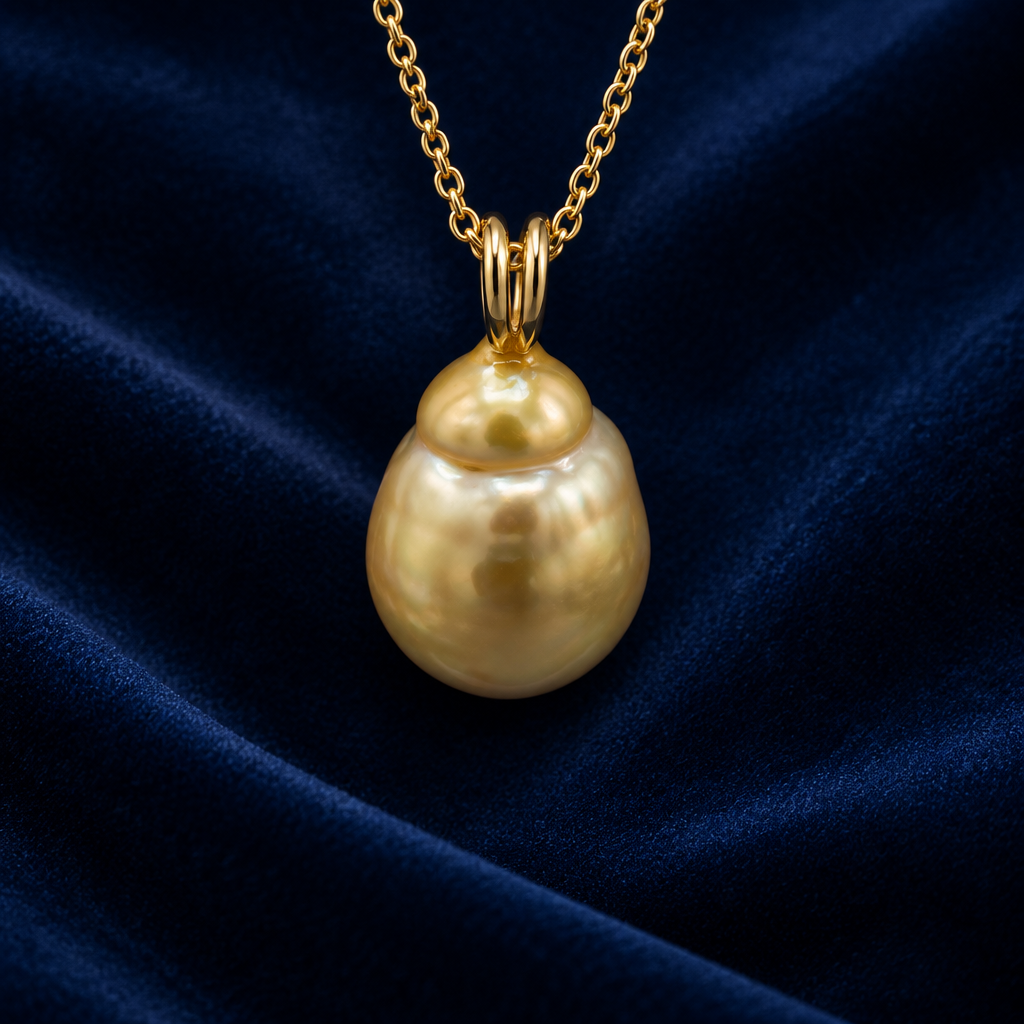 A gold pendant necklace featuring a large, baroque gold south sea saltwater pearl with a soft pearlescent luster, suspended from a delicate gold chain against a blue velvet background.