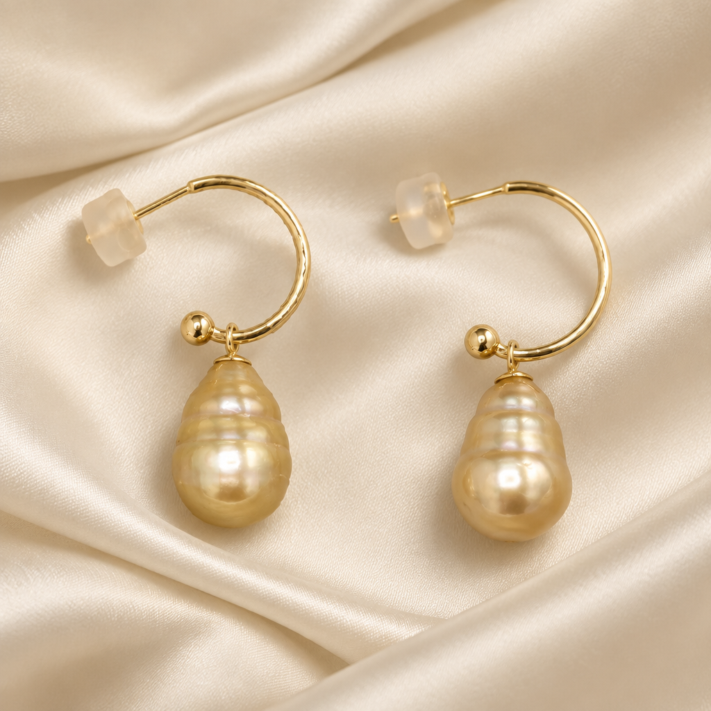 A pair of gold hoop earrings each with a large, baroque south sea golden saltwater pearl dangling from the front, displayed on a draped cream satin fabric background.