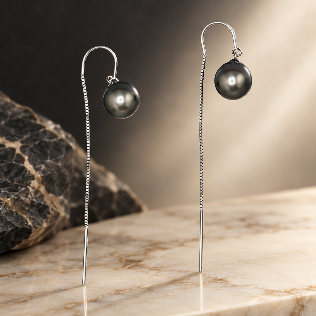 A pair of long drop earrings featuring dark gray Tahitian saltwater pearls suspended from white gold threaders with textured chain drops, set against a stone countertop background.