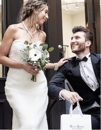 A bride in a strapless white wedding gown holding a white and green bouquet stands next to a smiling groom in a black tuxedo holding a white shopping bag with the brand name 'Holly Zhang'.