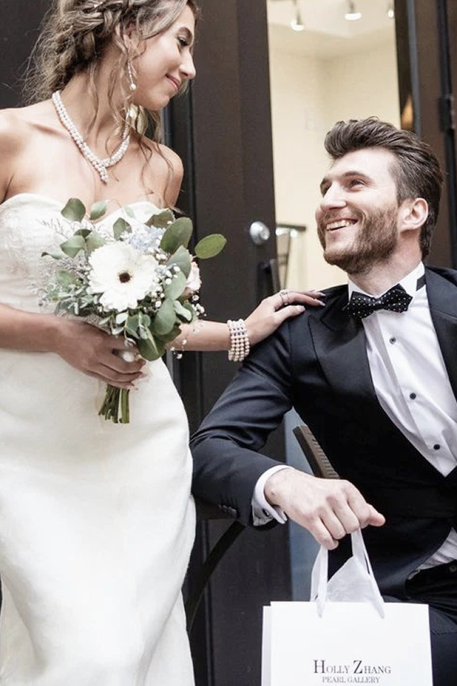 A model smiling and looking down, showcasing an elegant selection of pearl jewelry: a complex multi-strand pearl and gold necklace, delicate dangling pearl earrings, and a multi-row pearl bracelet, paired with a white strapless lace wedding dress and a floral bouquet.