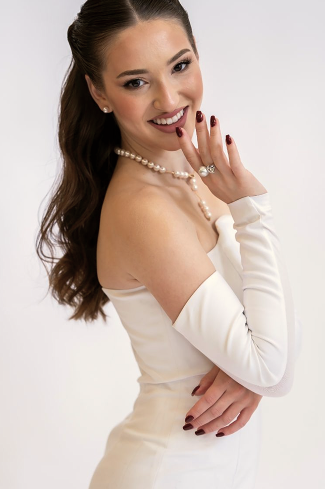 A model smiling, wearing an off-the-shoulder white top and detachable sleeves, showcasing an elegant jewelry collection featuring a multi-strand pearl and crystal necklace, small stud pearl earrings, and a large statement pearl and silver cocktail ring.