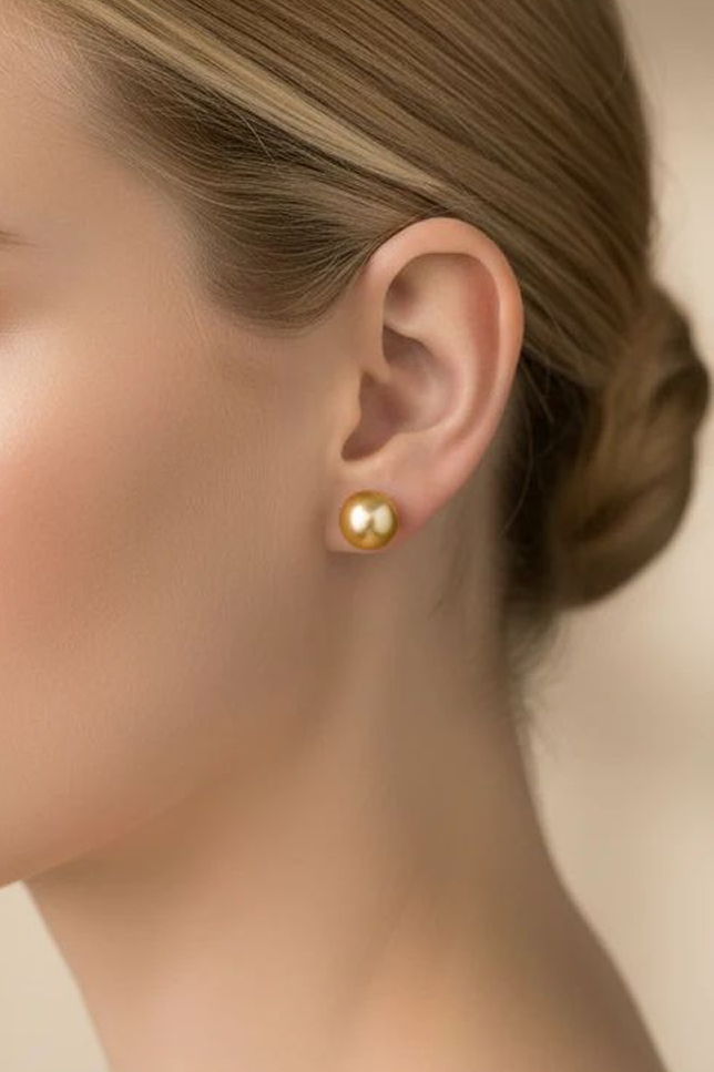 A model's left ear and neck, focusing on a single large, perfectly round golden South Sea pearl stud earring, set in a gold post and worn with her blonde hair tied up.