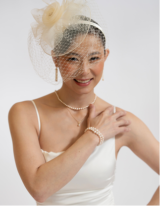 A woman wearing a white bridal-style dress and jewelry, including a pearl necklace, pearl bracelet, and a cream fascinator headpiece with a veil, against a light gray studio background.