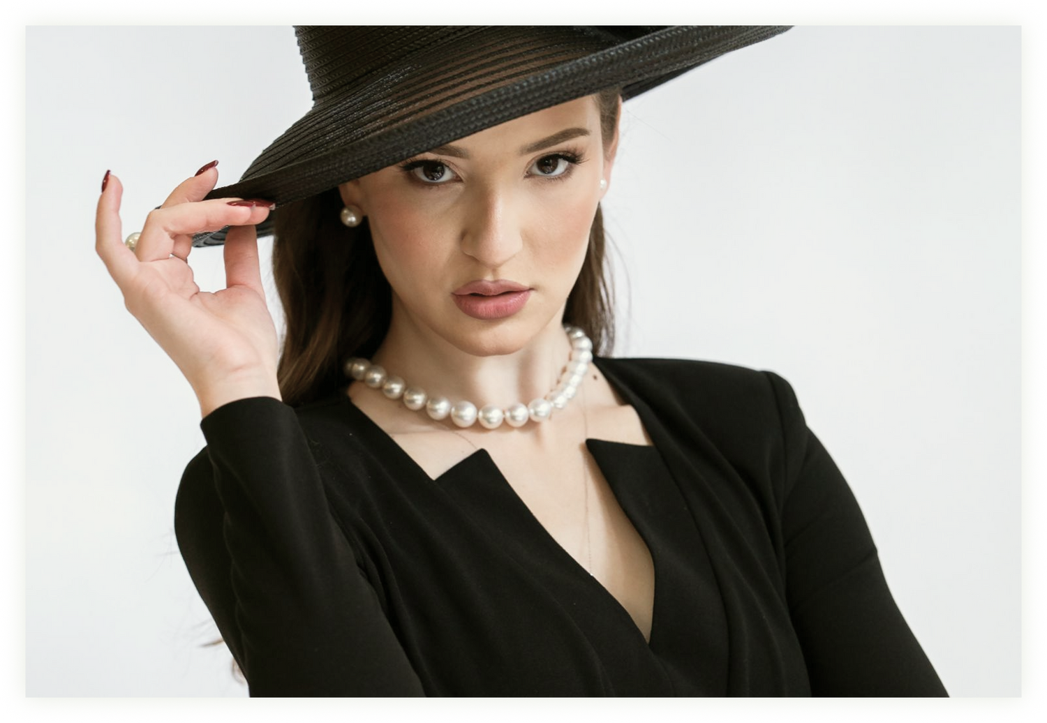 Woman wearing a white pearl necklace and matching pearl earrings with a black outfit and wide-brimmed black hat.