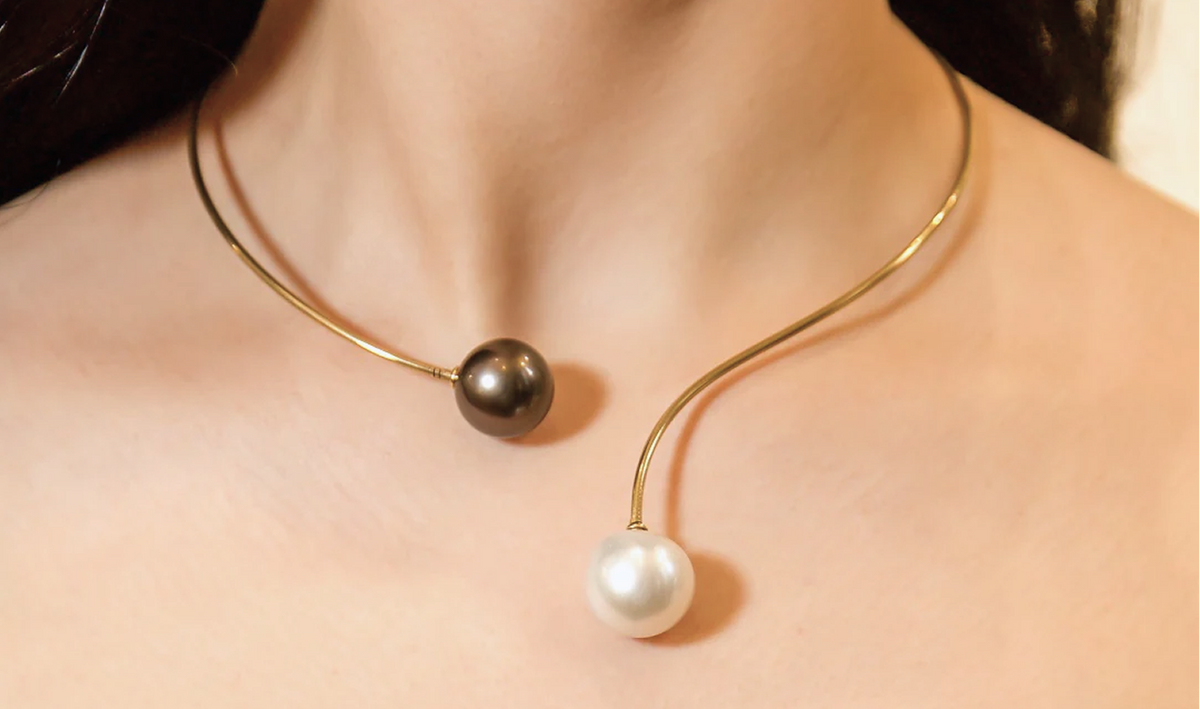 A close-up of a model's neck and chest wearing a delicate curved gold-tone necklace with two pearls, one dark charcoal and one white, on a soft beige background.