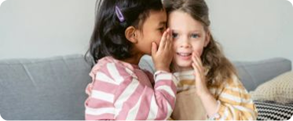 Two young girls sitting on a grey couch whispering a secret to each other.