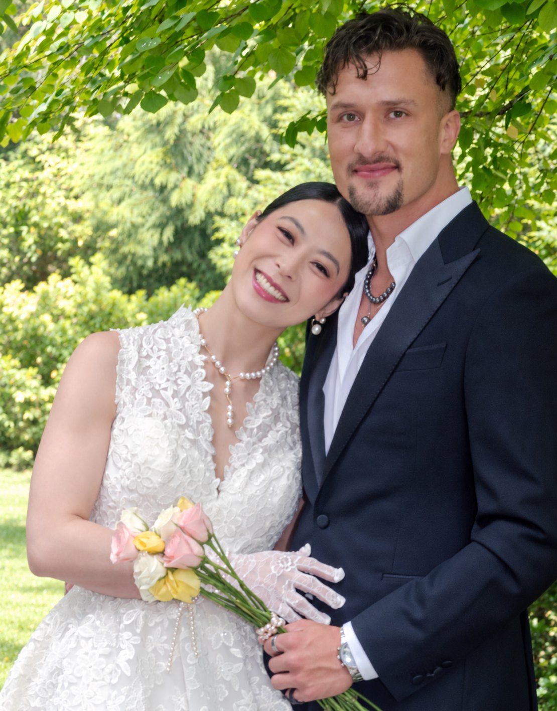 A bride and groom smiling together outdoors, the bride wearing pearl earrings and a pearl necklace