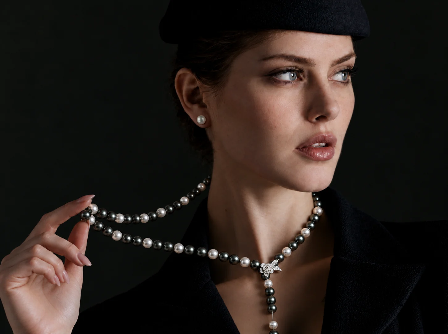 A model in a black beret and jacket against a dark ground, holding up a long necklace of Tahitian and white pearls with a pavé flower clasp, wearing a white pearl stud