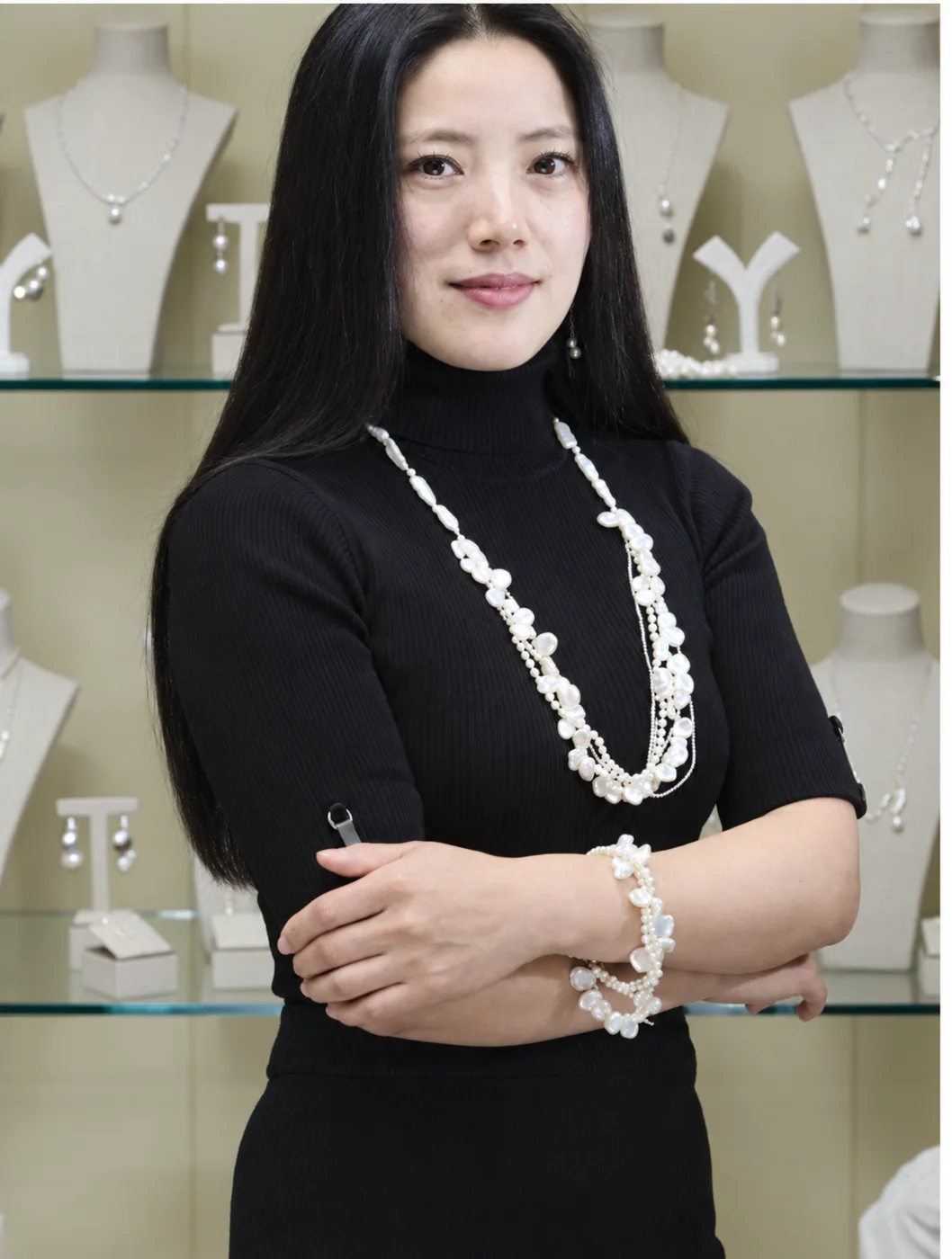 Holly Zhang in a black turtleneck, arms folded, wearing a long keshi pearl necklace and bracelet in front of the gallery displays
