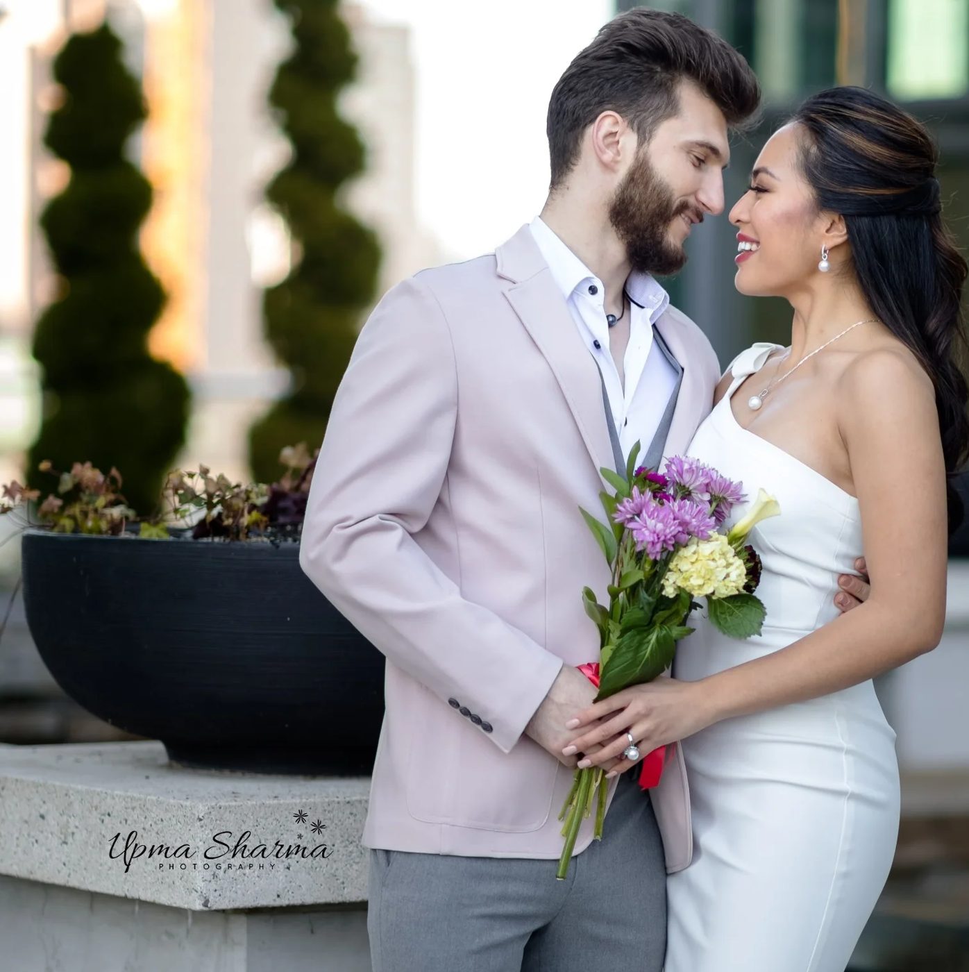 A bride and groom outside, she in white with a pearl necklace and a bouquet